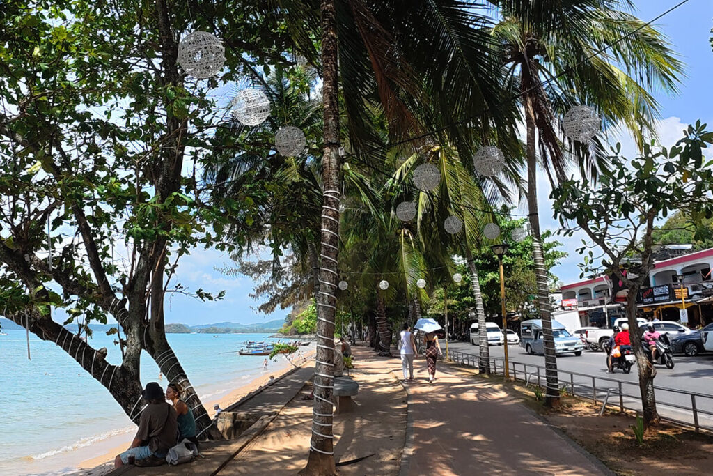 Wide seafront promenade in Ao Nang with hanging lanterns on coconut palms and coastal road alongside