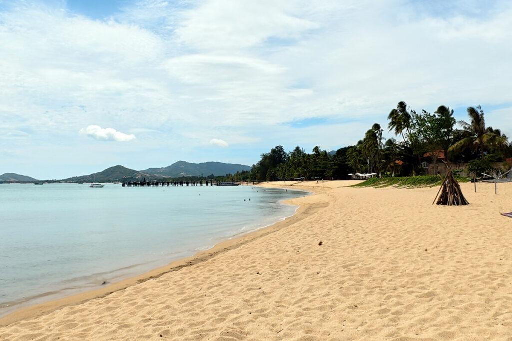 Wide golden sand beach in Maenam Koh Samui with lush green hills and wooden pier pilings in the distance