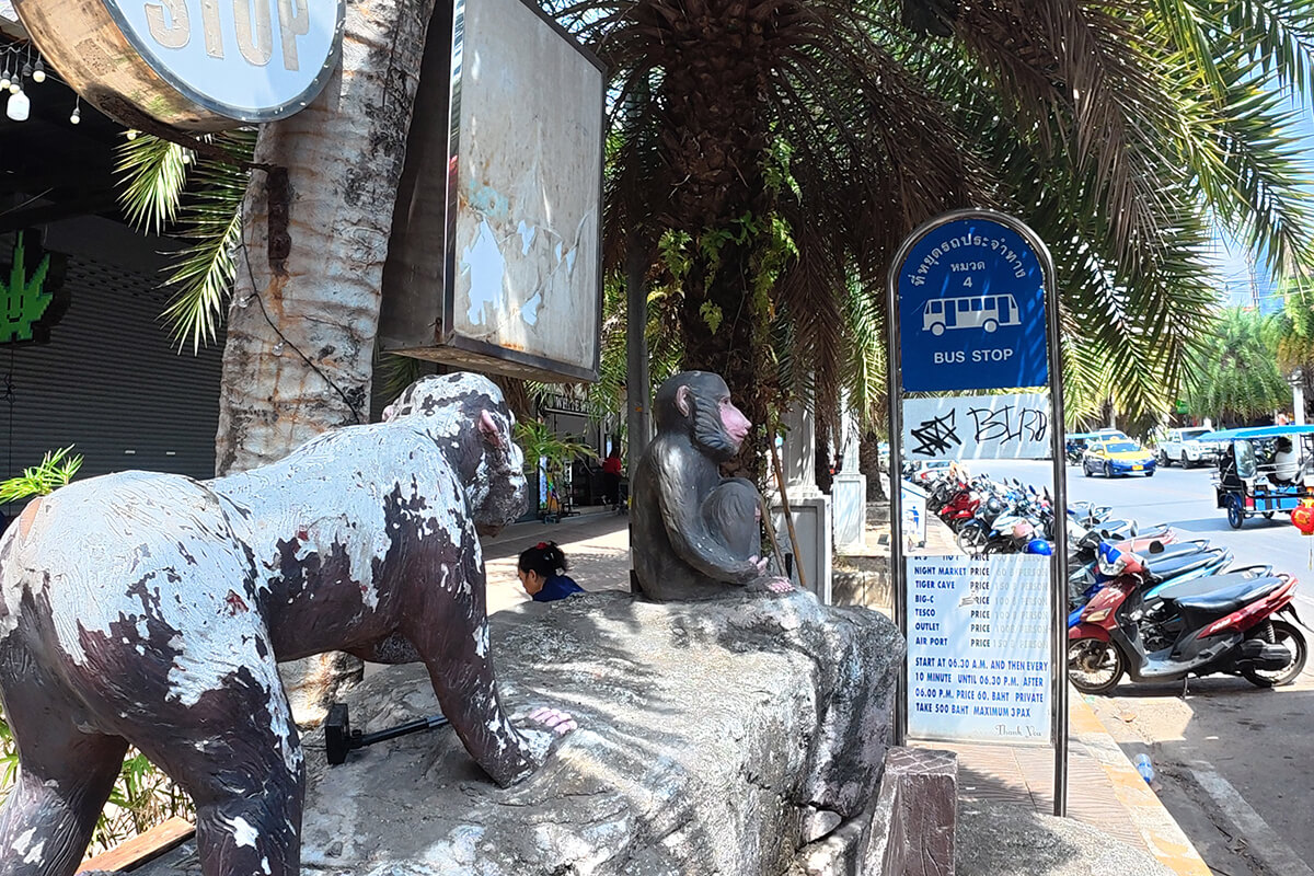 Blue local bus stop timetable sign beside painted monkey statues along the pavement in Ao Nang, Krabi