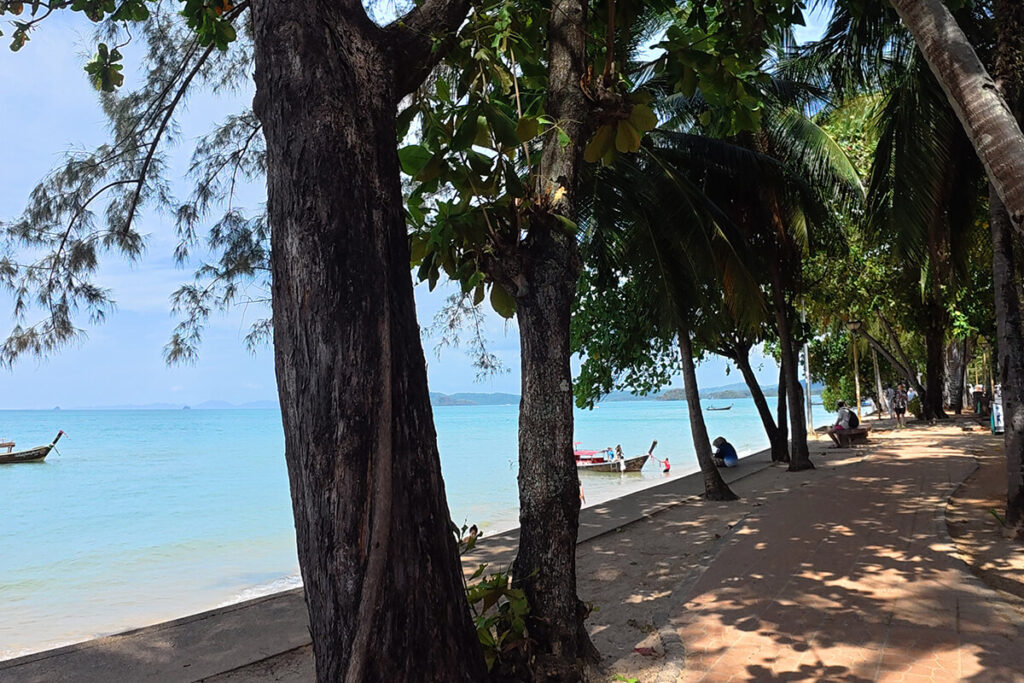 Paved footpath under coastal trees with longtail boats floating in the sea at Ao Nang Beach