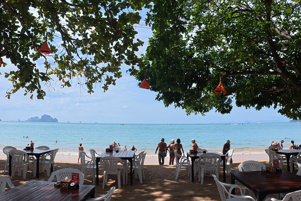 Dining tables set directly on the sand shaded by mature tropical trees looking out over the sea at The Last Fisherman Bar